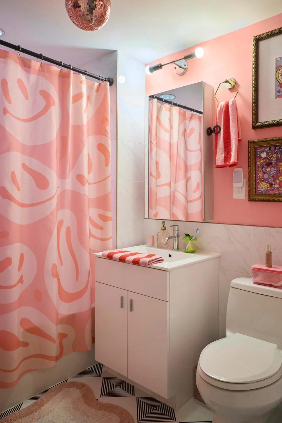 A pink patterned shower curtain, coral hand towel, and folded striped towel surround a small white vanity.