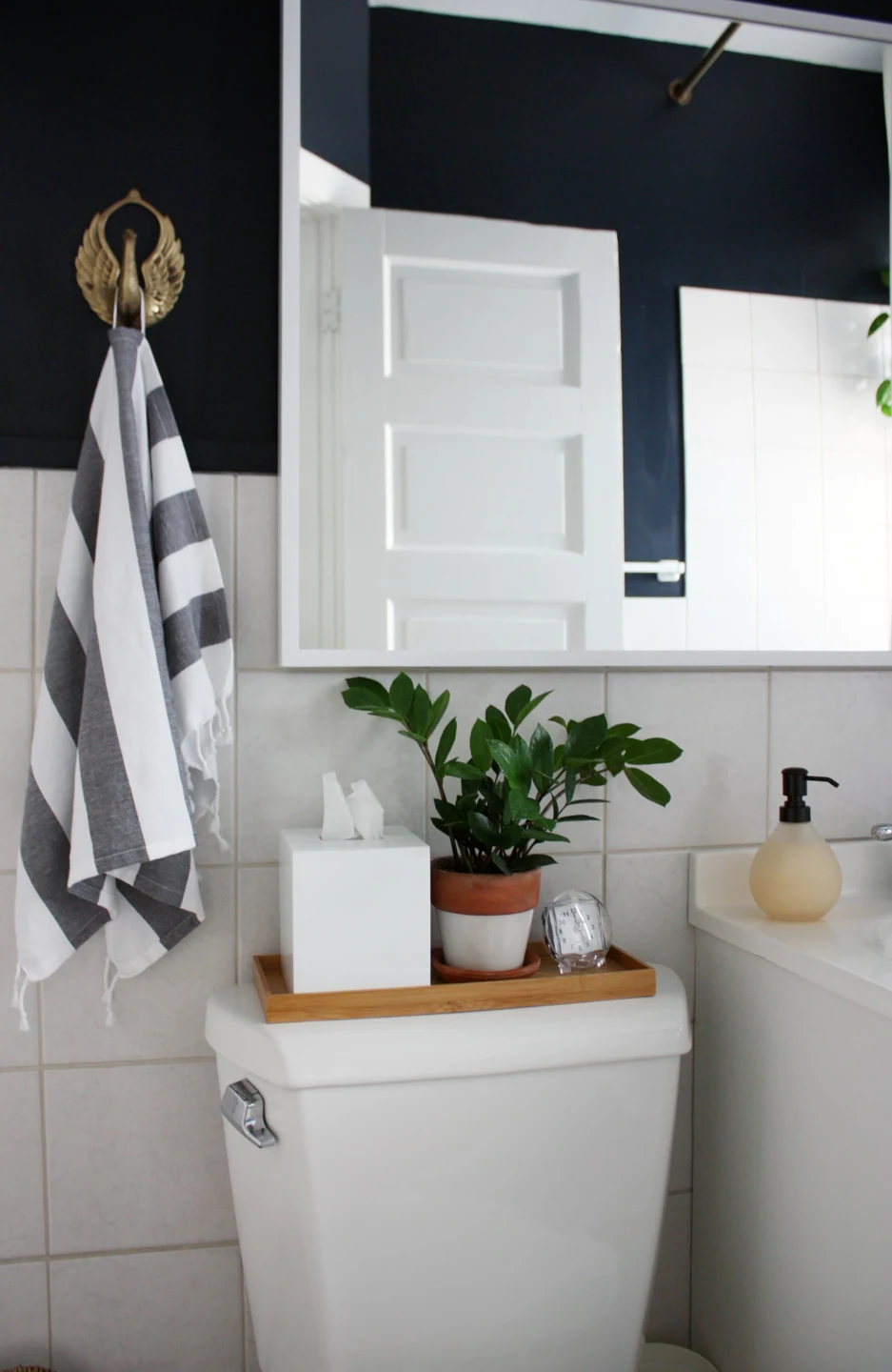 A narrow wood-toned tray holds a white tissue box, a potted plant, and a small clock on a toilet tank.