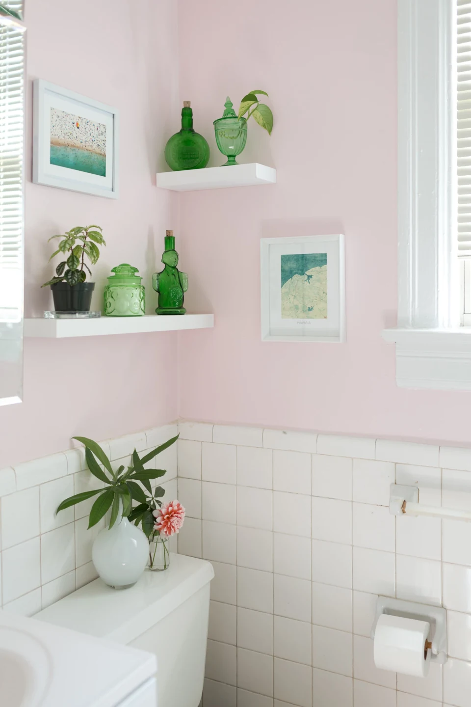 Green bottles and a lidded jar sit on white bathroom shelves above a white vase and a small flower in a clear bottle.