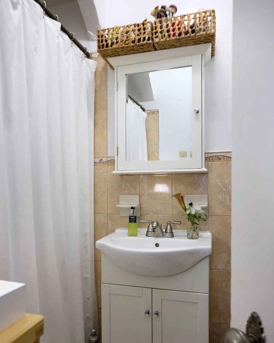 Two open woven baskets hold brushes and small bottles above a mirrored cabinet, with a small flower vase beside the sink.