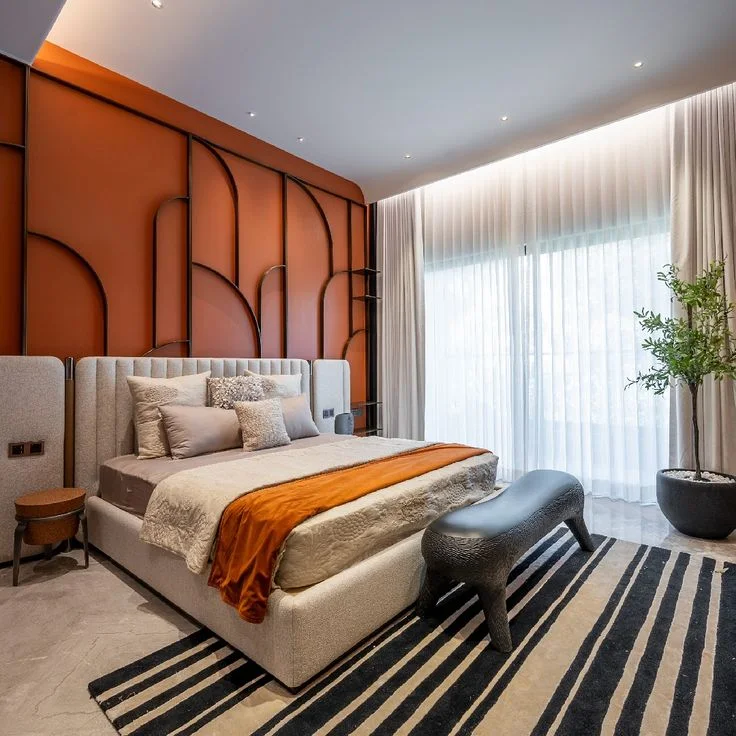 An orange accent wall with dark curved lines stands behind a pale bed and an orange throw.