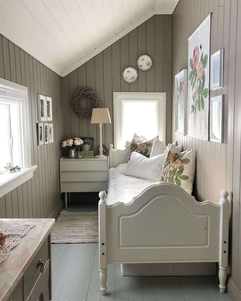 A tall pale table lamp stands on a small chest beside a white bed in a paneled bedroom.