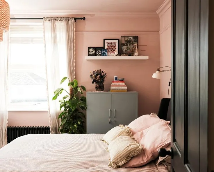 Pink walls and pink pillows sit beside white bedding, cream curtains and a gray-green cabinet.
