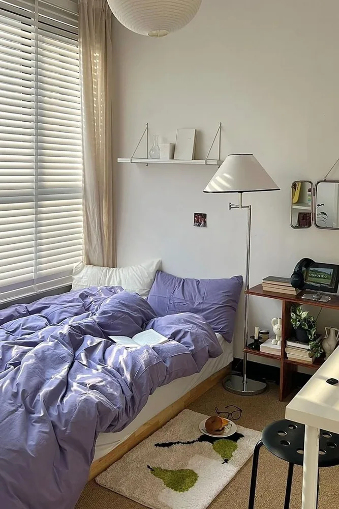 White bedroom walls sit behind lavender bedding, a floor lamp and a small open bedside shelf.