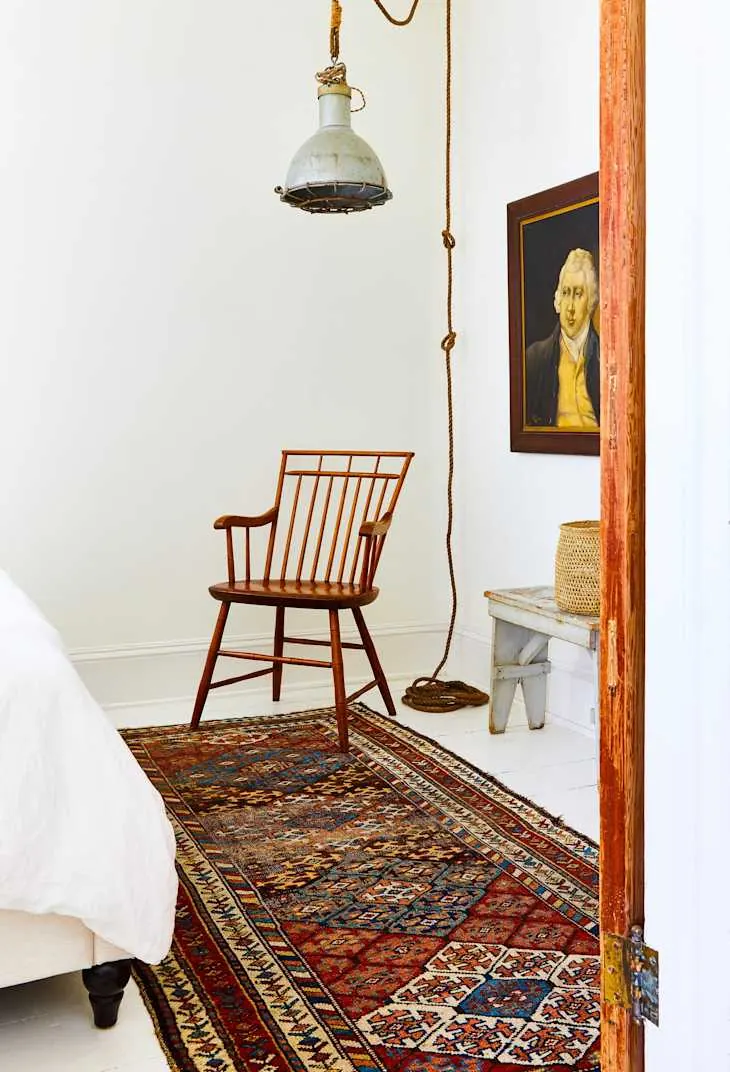 A colorful patterned runner sits near the foot corner of a white bed beside a wooden chair.