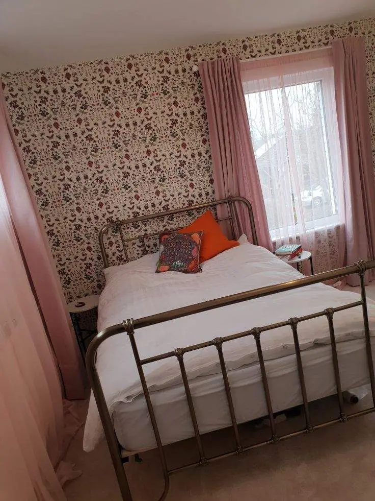 Plain pink curtains and white sheers beside floral wallpaper, white bedding, and a metal bed frame.