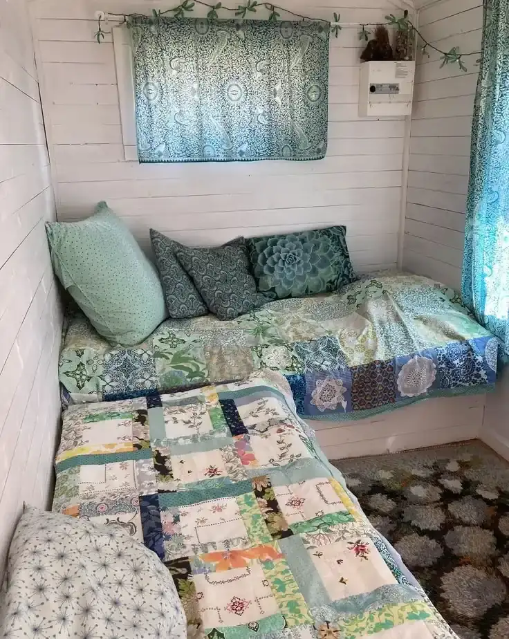 Two narrow daybeds have pale patchwork covers and blue-green pillows beside patterned curtains.
