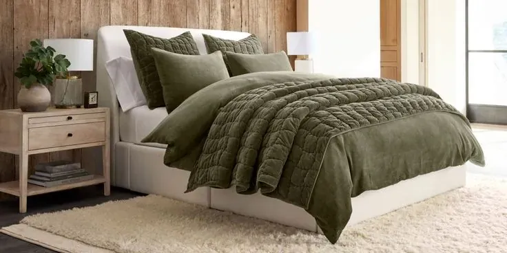 An olive quilt covers a pale bed above a cream rug beside wood-toned nightstands.