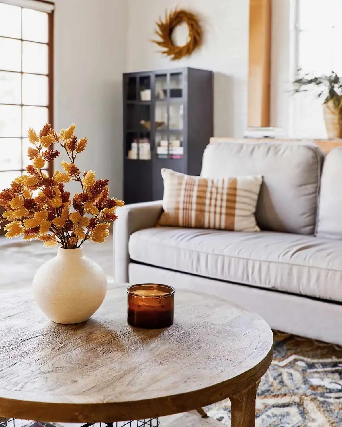 A cream and brown plaid pillow sits on a gray sofa behind a vase of golden leaves.