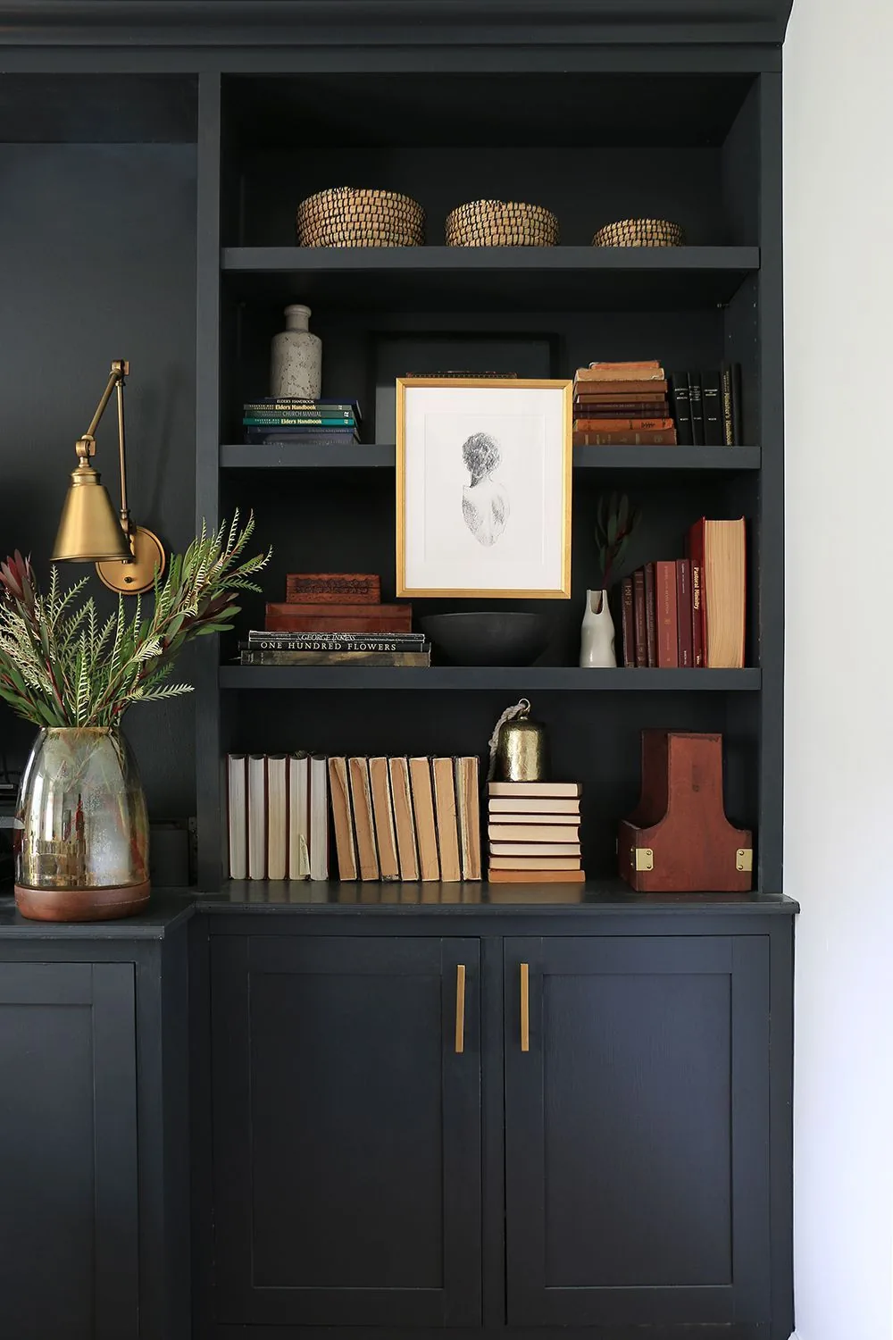 Earth-toned books, woven baskets, a framed drawing, and small vessels fill dark built-in shelves.