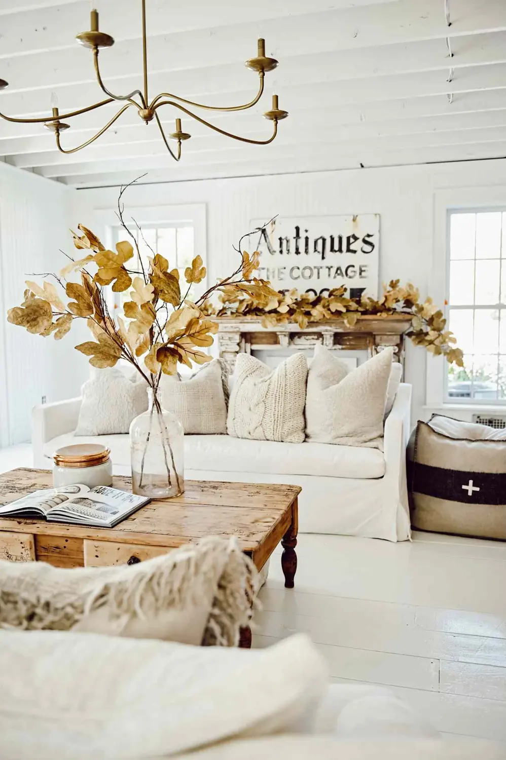 Golden leaves in a clear vase stand in front of a cream sofa and a mantel draped with more leaves.