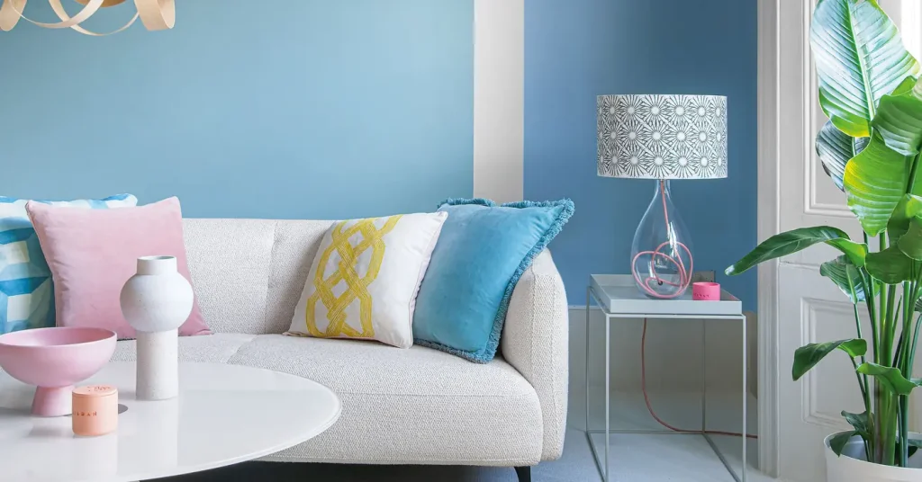 A white sofa with pink, blue, and yellow cushions sits against a blue wall beside a lamp and broad green leaves.