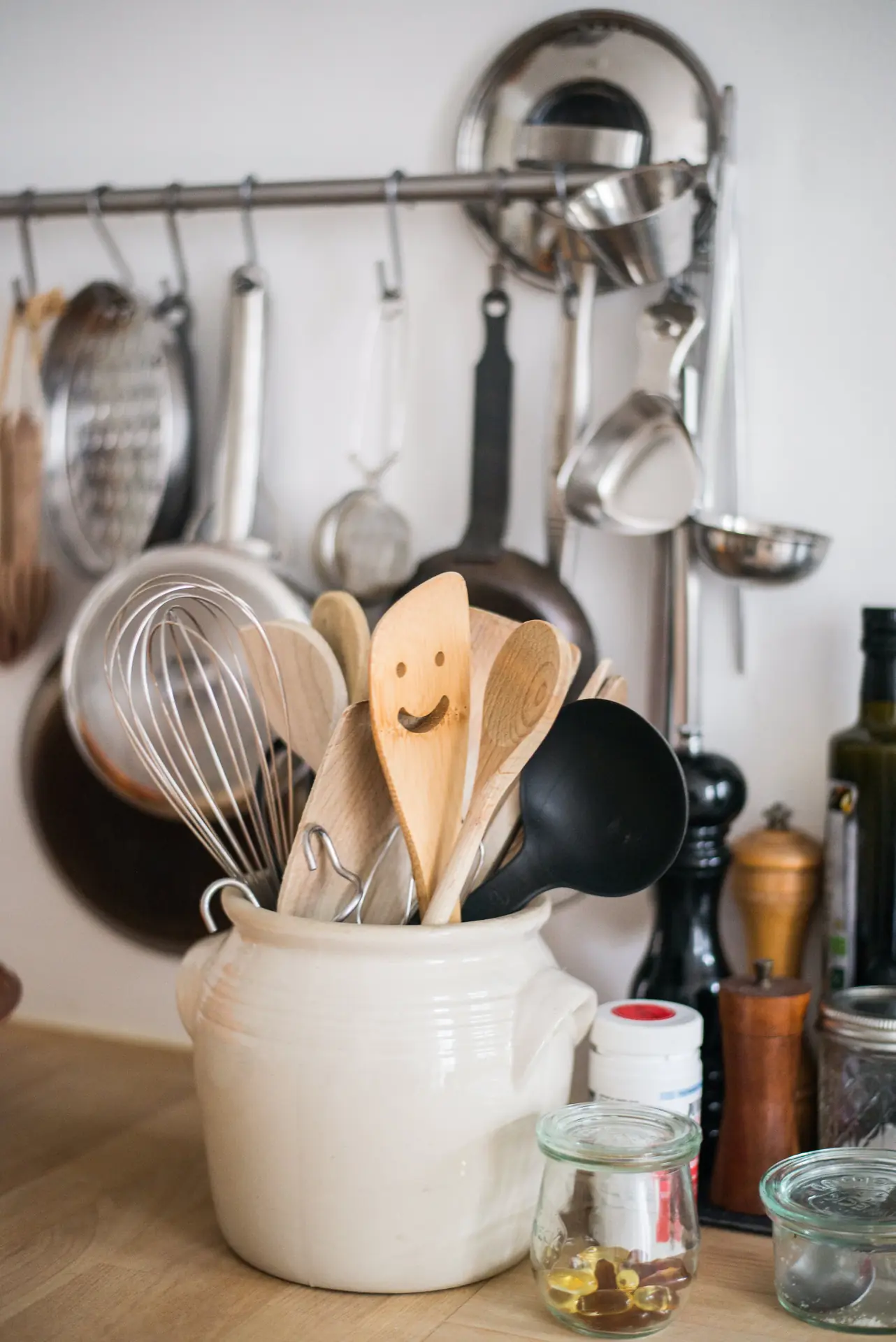A white utensil crock holds wooden spoons, a whisk, and a black ladle on a kitchen counter.