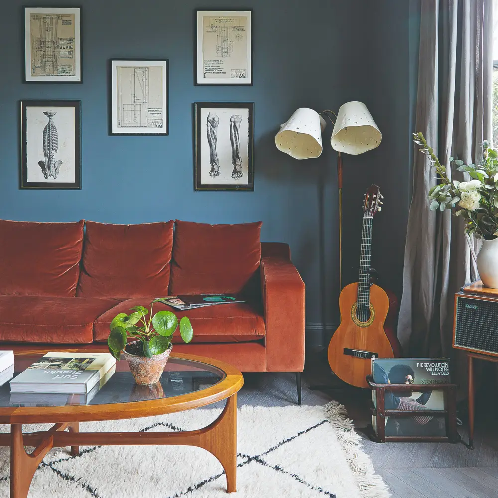 A muted blue wall stands behind a rust sofa and a white rug with dark lines.