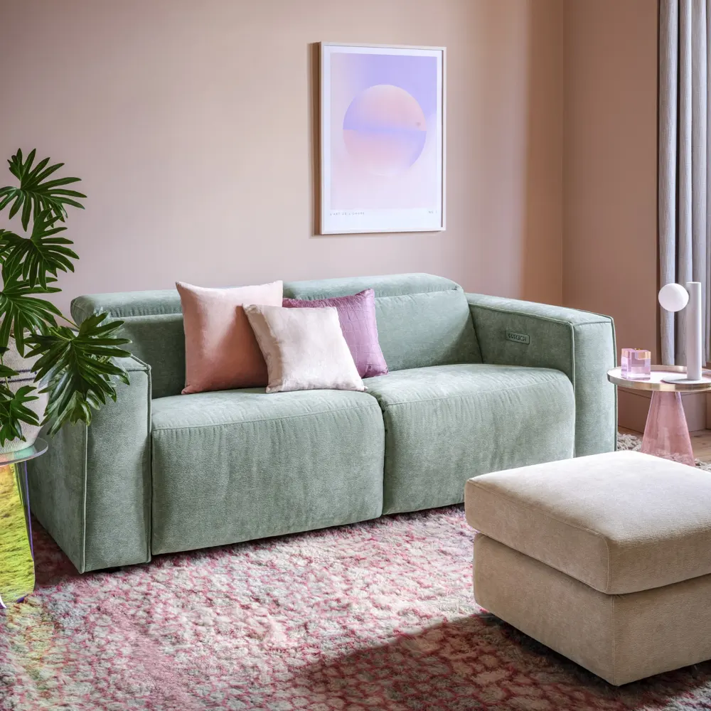 A pale pink wall sits behind a muted green sofa with pink cushions and a patterned rug.