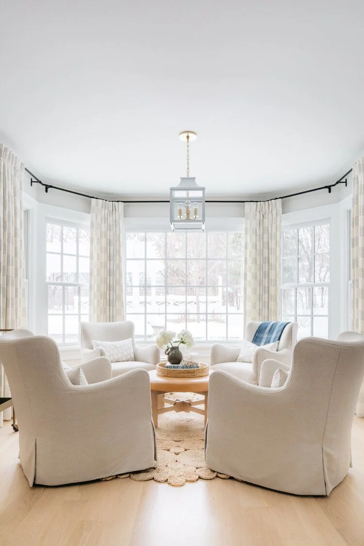 Pale patterned curtain panels follow a bay window around four cream armchairs.