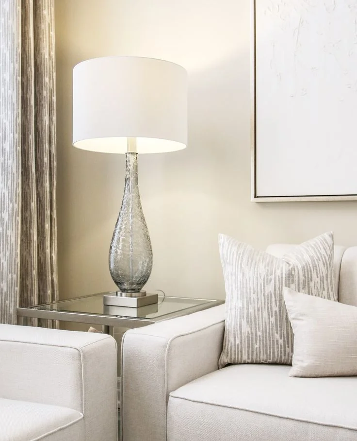 Cream seating with pale striped cushions stands beside a glass table and a lamp with a broad white shade.