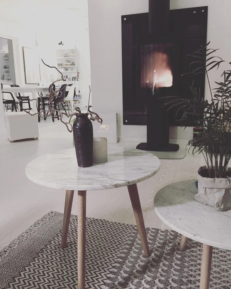 Pale round tables and a dark vase stand before a black fireplace above a patterned rug.