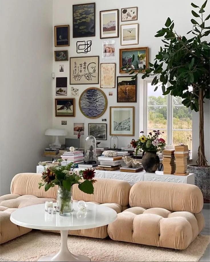 A tall gallery of mixed artwork rises behind a pale sculptural sofa beside a window.