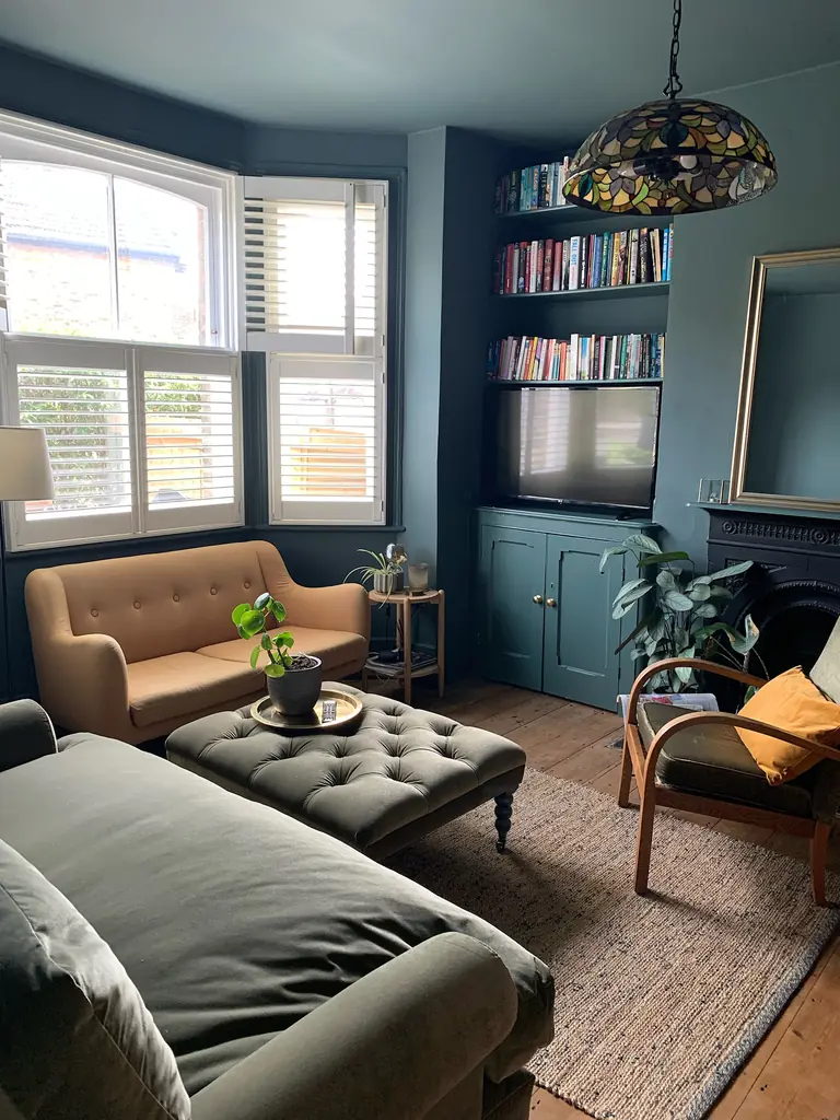 A tufted ottoman sits between a sofa, a loveseat under the window, and a wooden armchair in a dark blue living room.
