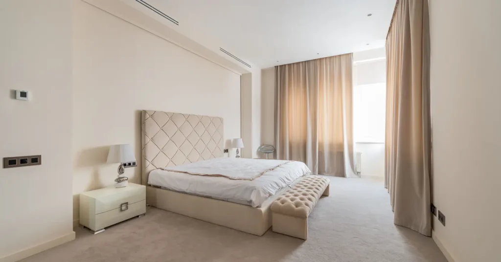 Neutral bedroom with a cream upholstered bed, white bedding, a tufted bench, and beige curtains.