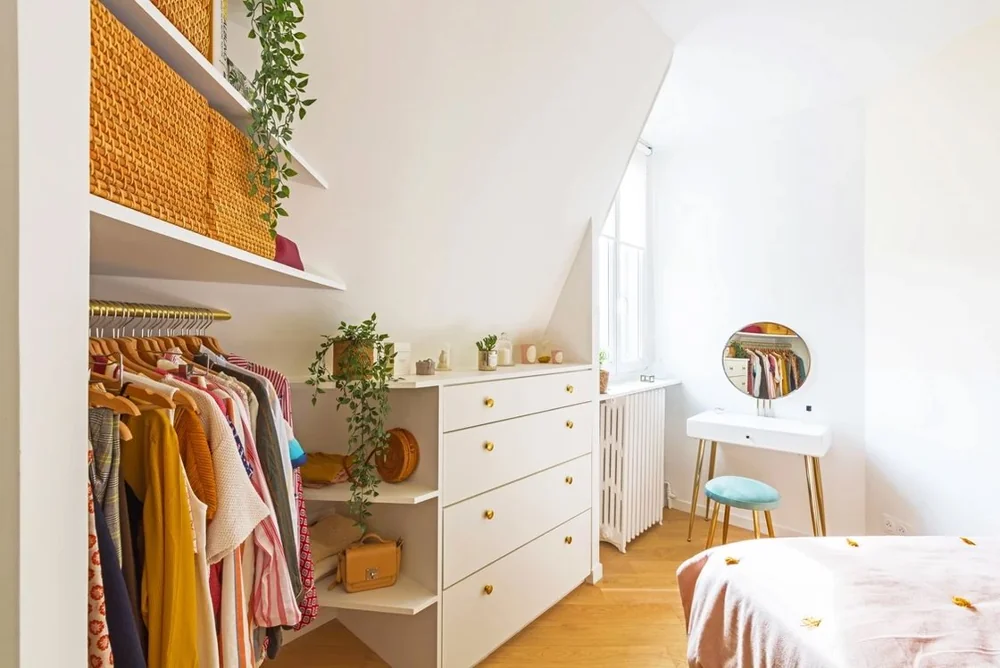 Open clothes storage and a chest of drawers face a bed under a sloping ceiling.