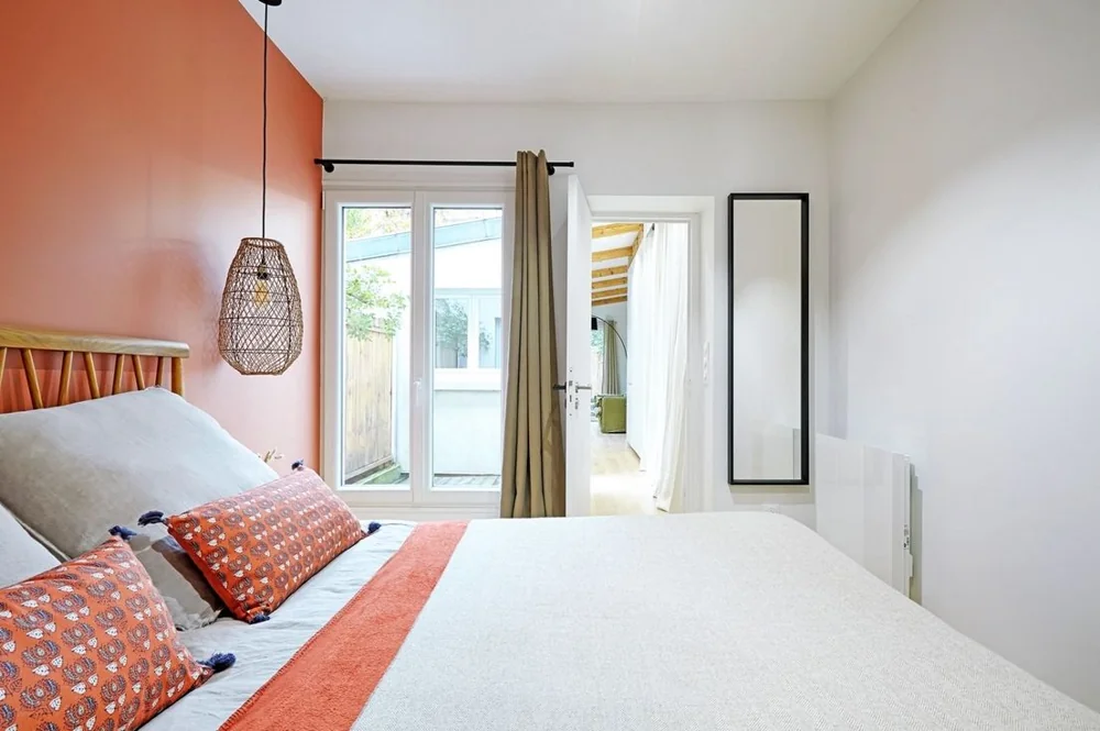 A bed sits against a coral side wall beside a window and a tall mirror.