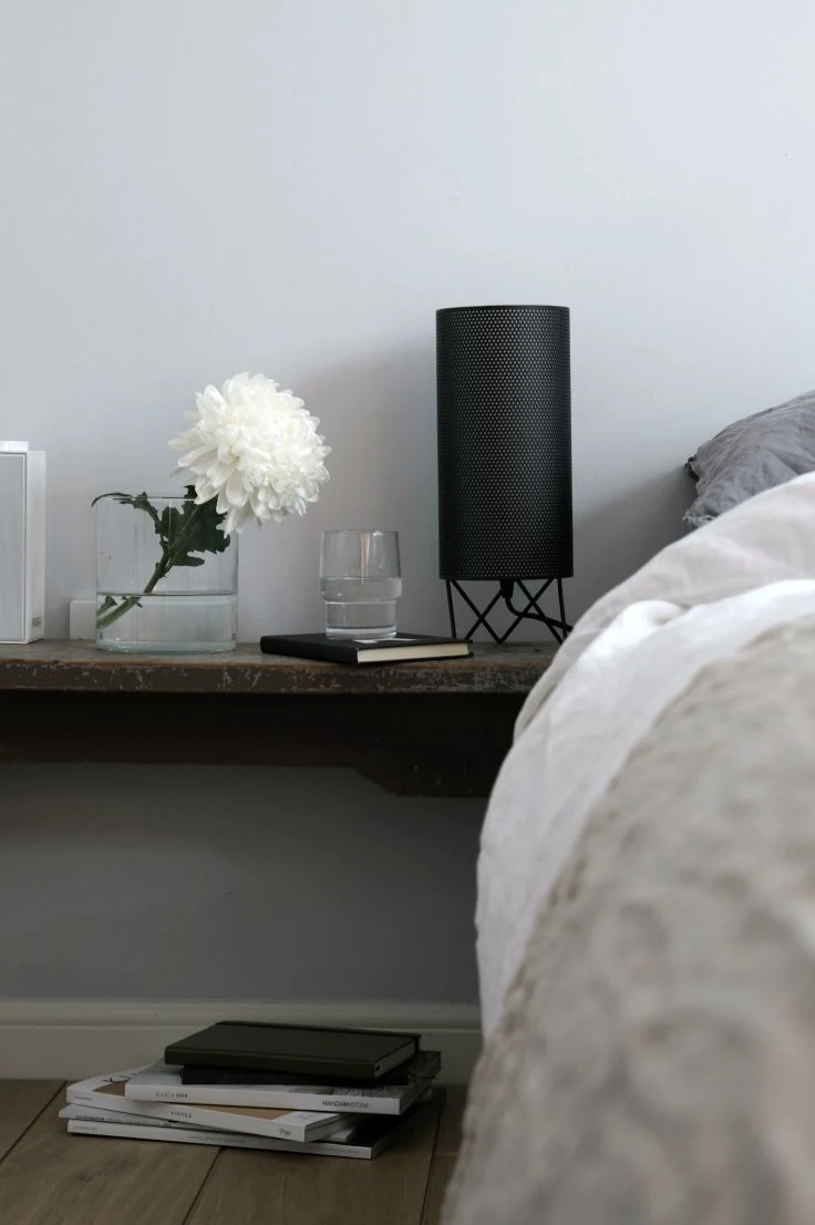 A narrow bedside shelf with a black lamp, white flowers, and a glass.