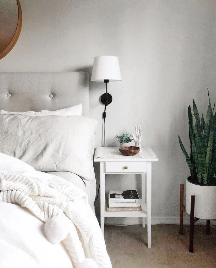 A small white nightstand beside white bedding and a wall-mounted lamp.