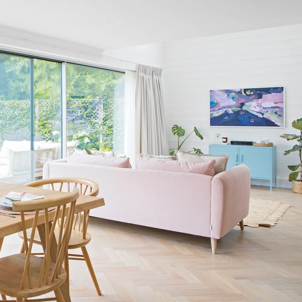 A pink sofa separates a dining table from a living area beside wide glazed doors.