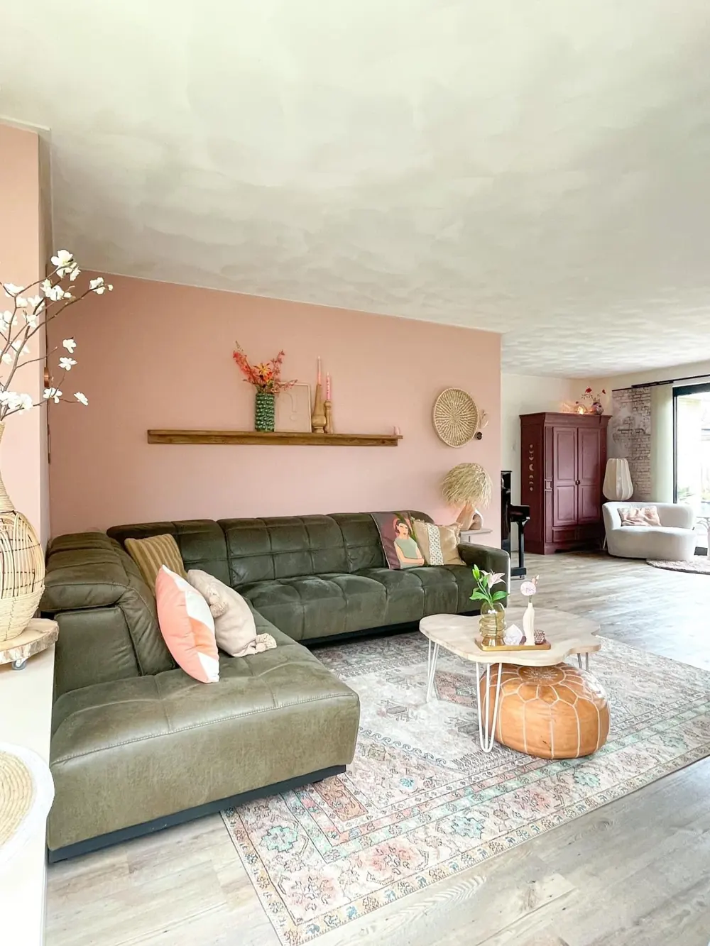 An olive sectional with pink pillows, a pink wall and a pale patterned rug.