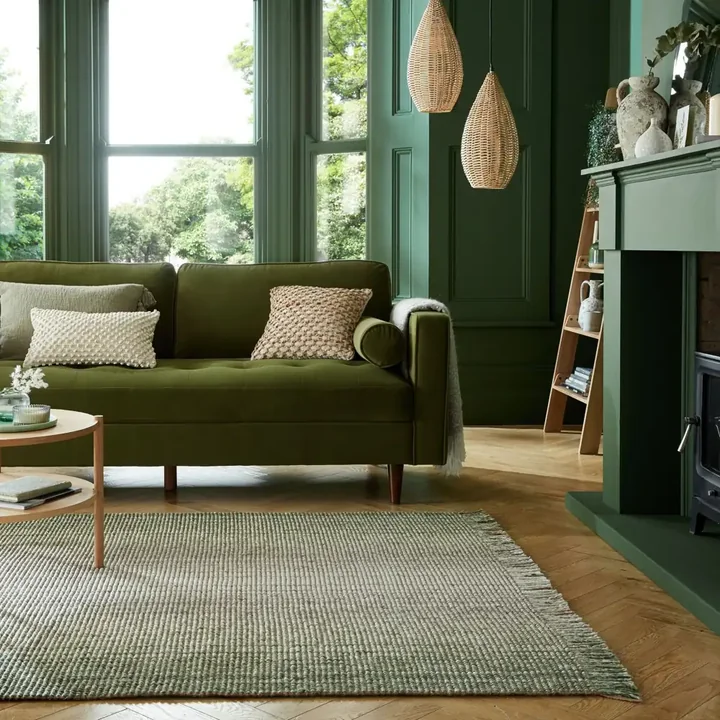An olive green sofa with cream pillows beside dark green walls and a pale green rug.