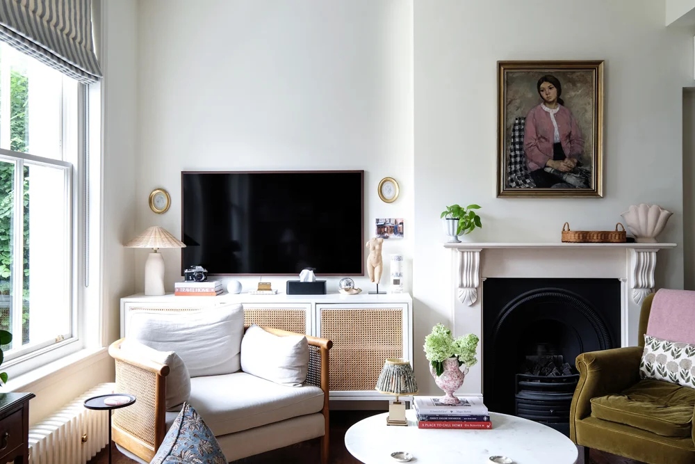 A light armchair stands beside a small round side table near a bright window and fireplace.